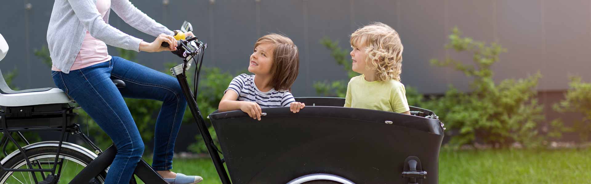 Eine Frau fährt mit einem Cargobike, in der Transportbox befinden sich 2 Kinder